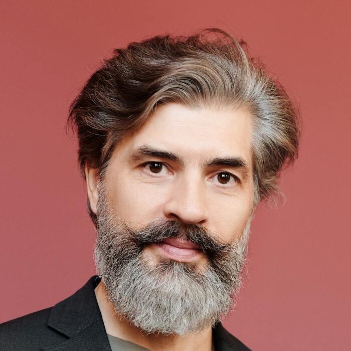 Portrait of a stylish bearded man holding eyeglasses against a pink background.